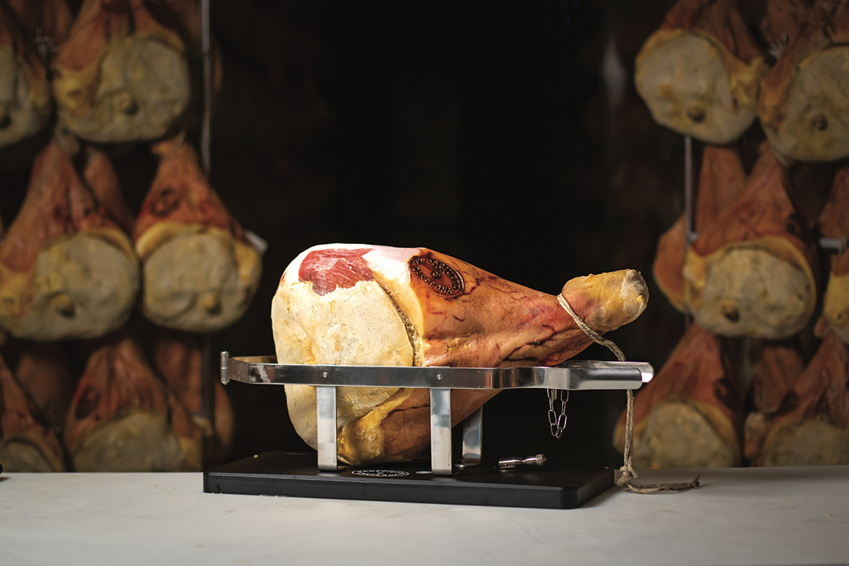 Prosciutto di Carpegna DOP su supporto. Salumi tipici italiani in stagionatura sullo sfondo.