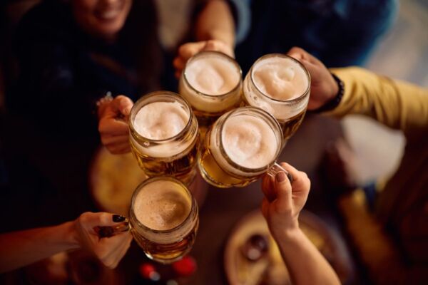 Brindisi con birra AssoBirra. Gruppo di amici celebra con pinte di birra chiara. Nuovi rituali e consumi consapevoli.