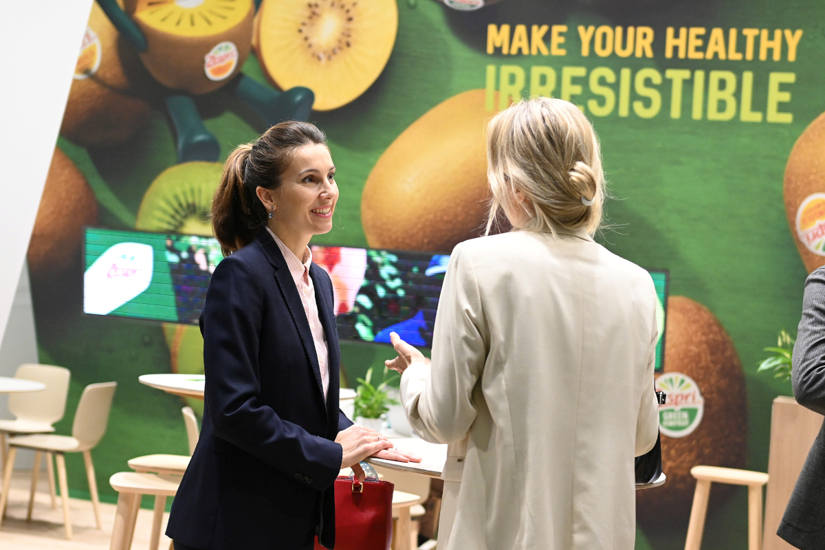 Due donne a Fruit Logistica. Stand con kiwi Zespri e slogan Make Your Healthy Irresistible. Fiera ortofrutticola.