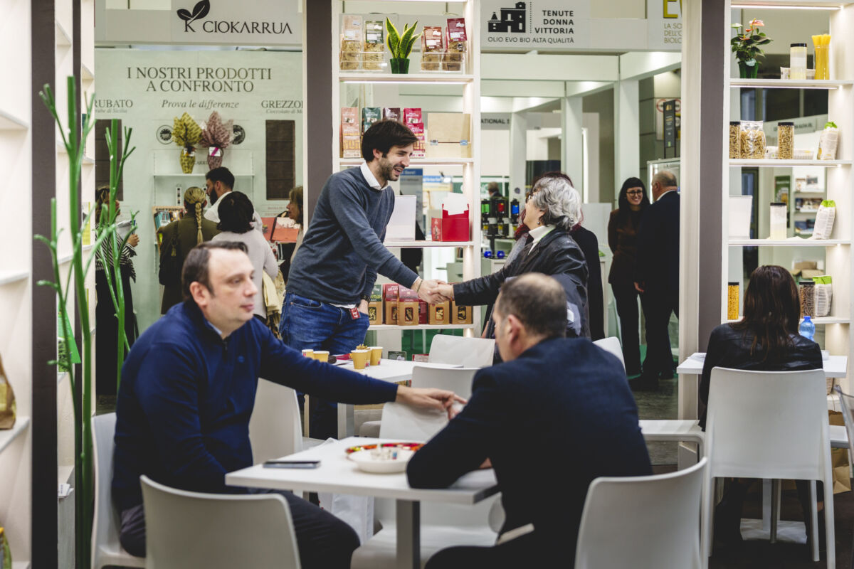 Stand prodotti alimentari SANA: stretta di mano, tavoli con persone sedute. Prodotti bio e naturali sullo sfondo.