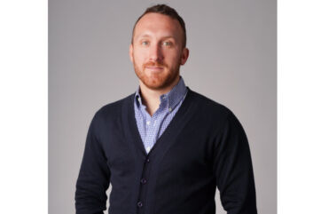 Rom'antica - Ritratto di Michele Mannara. Uomo caucasico con barba rossa, indossa camicia a quadri e cardigan blu. Sfondo grigio.