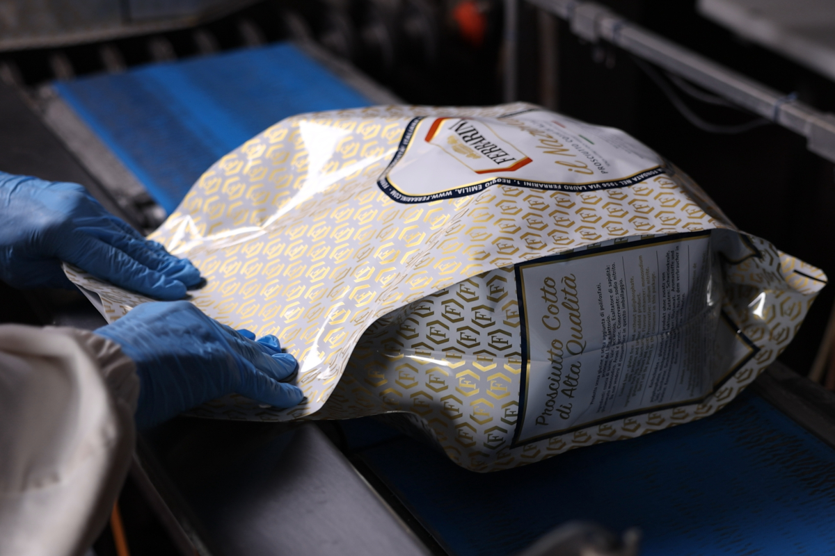 Lavorazione prosciutto cotto Ferrarini. Mani con guanti blu confezionano il prodotto su nastro trasportatore.