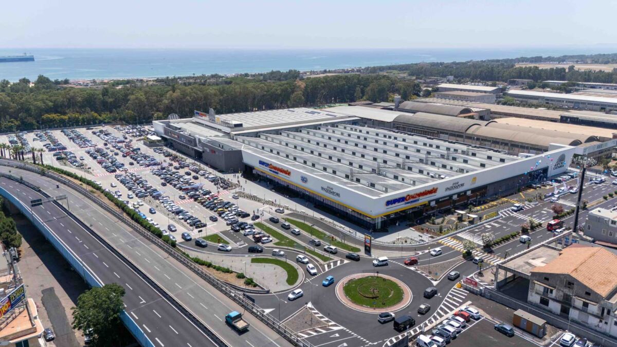 Veduta aerea de L'Arena di Catania, hub commerciale sostenibile con ampio parcheggio e viabilità vicino al mare.