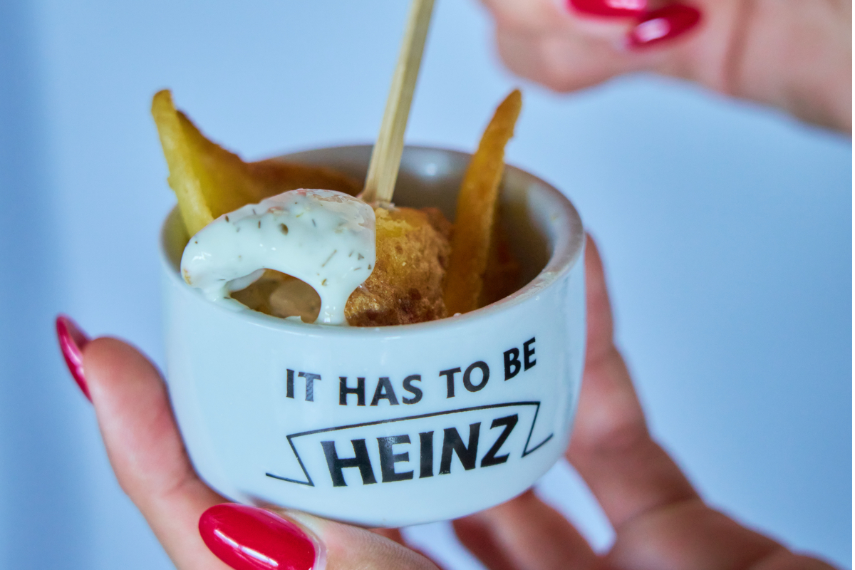 Mano con smalto rosso regge una ciotolina Heinz con patatine fritte e una delle nuove salse Heinz per il mercato italiano.
