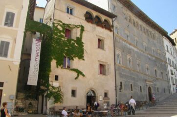 Facciata di Casa Menotti a Spoleto, edificio storico con edera e tavolini all'aperto in una piazza soleggiata.