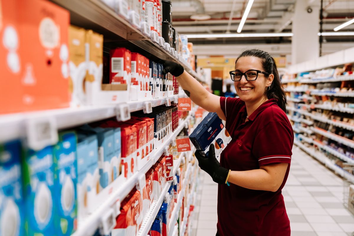 Dipendente Nova Coop sorridente riordina gli scaffali del supermercato con prodotti a marchio Coop.