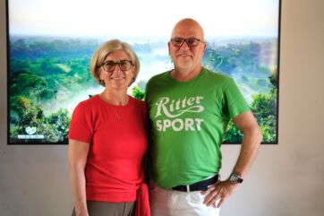 Due persone sorridenti, di cui una con maglietta Ritter Sport, davanti a uno sfondo di foresta per il cioccolato sostenibile.