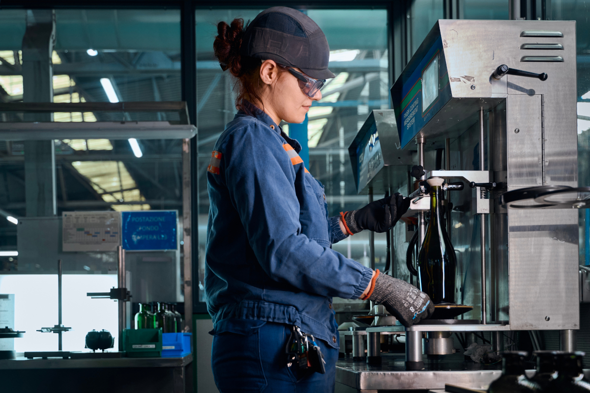 Operaia Verallia con DPI lavora in sicurezza su un macchinario industriale per il controllo qualità di bottiglie in vetro.