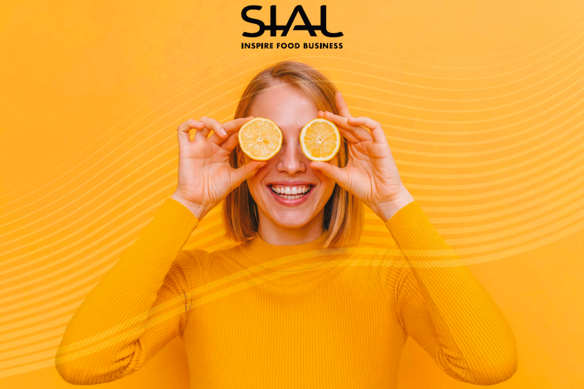 Donna sorridente con fette di limone sugli occhi su sfondo giallo, logo SIAL Paris 2026 per le tendenze alimentari.