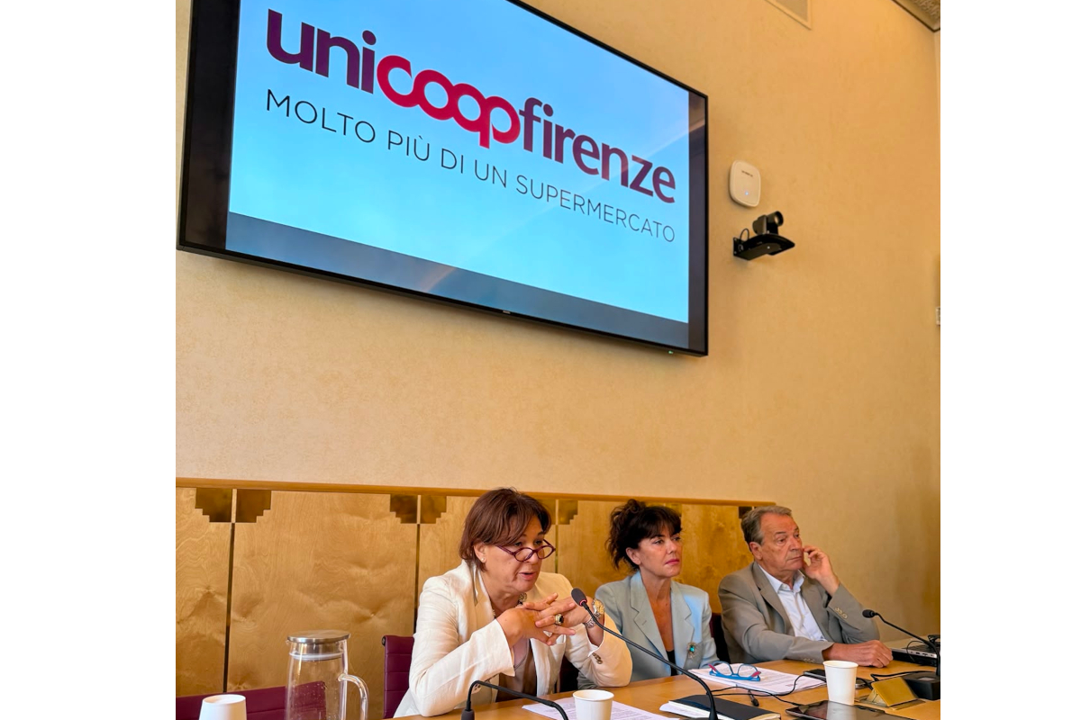 Conferenza stampa Unicoop Firenze: relatori al tavolo con il logo aziendale proiettato sullo schermo alle spalle.