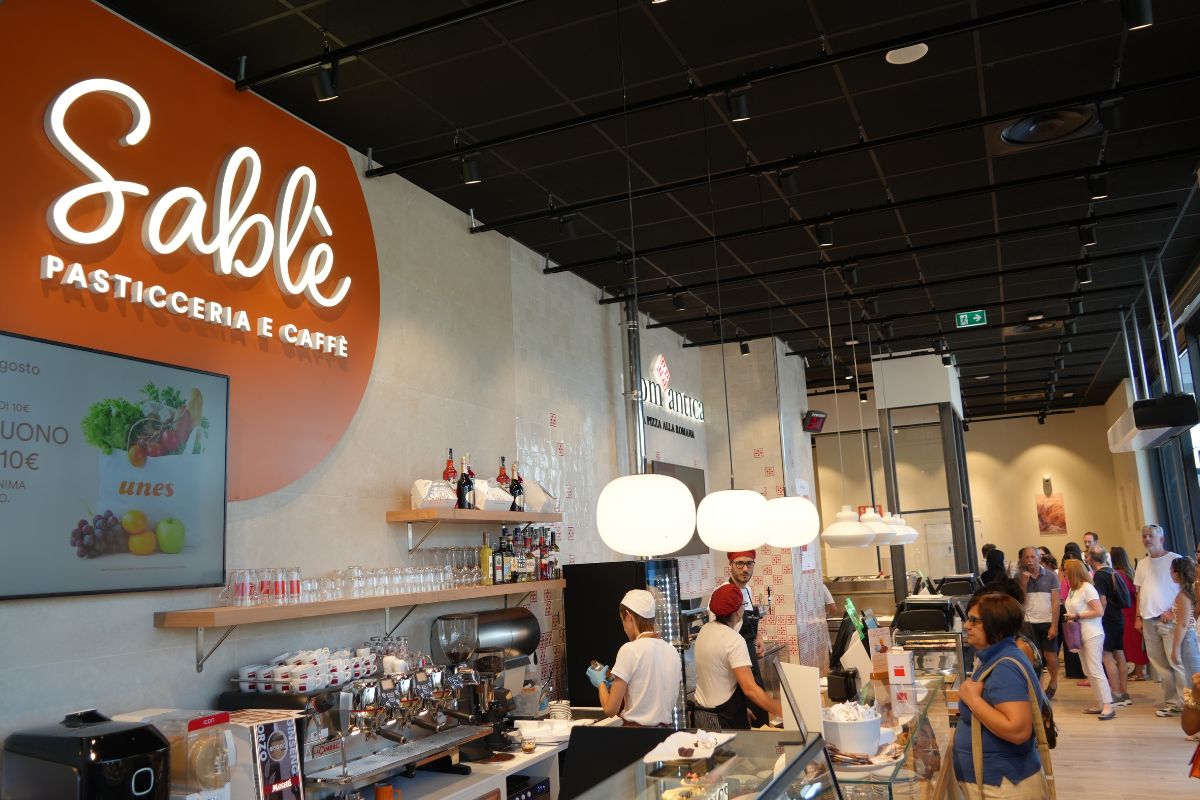 Interno del nuovo concept store Unes con pasticceria e caffè Sablé, bancone bar, personale al lavoro e clienti.
