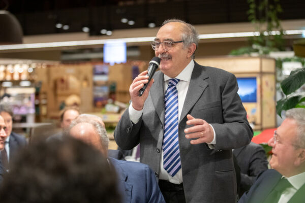 Paolo parla al microfono durante un evento conviviale a Fico, circondato da ospiti in giacca e cravatta.