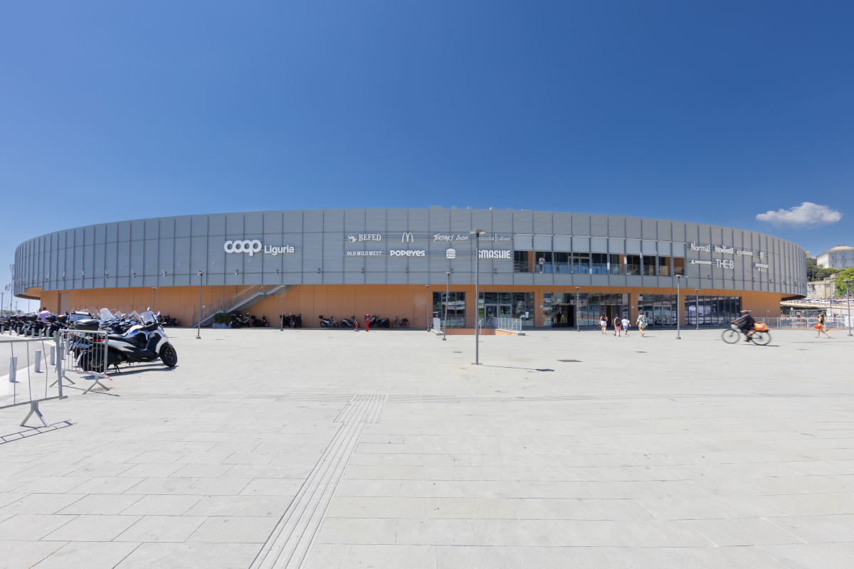 Facciata esterna del nuovo Coop Waterfront di Genova con insegne retail e illuminazione Imoon in una piazza soleggiata.