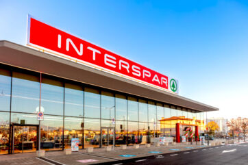 Facciata esterna di un supermercato Interspar Despar Nord con vetrate ampie e insegna rossa luminosa in una giornata soleggiata.