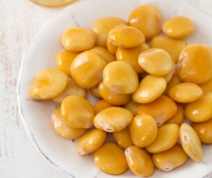Piatto di lupini salati serviti come snack healthy insieme a un bicchiere di birra fresca su tavolo bianco.