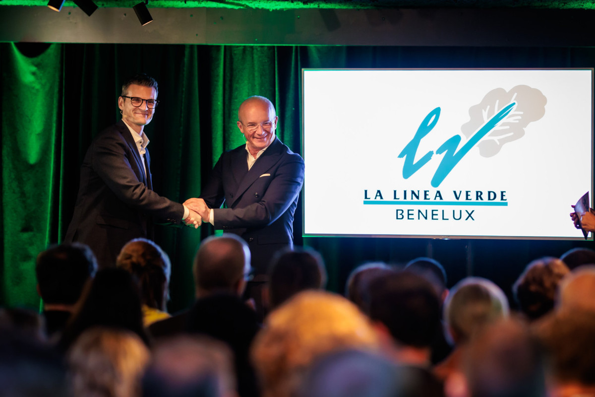 Stretta di mano tra dirigenti sul palco durante la presentazione di La Linea Verde Benelux, logo aziendale sullo schermo.