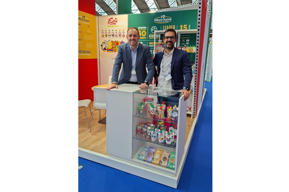 Due uomini sorridenti allo stand Natura Nuova alla fiera, con prodotti esposti e insegne aziendali.