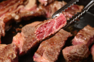 Pinza che solleva una fetta di carne bovina al sangue durante la cottura in padella, focus su qualità della carne.