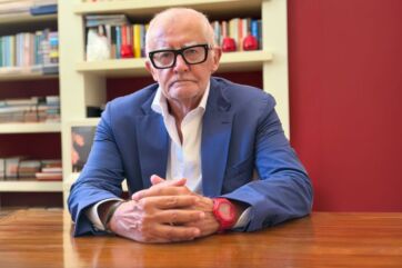 Mario Gasbarrino con occhiali neri e giacca blu, seduto al tavolo con libri sullo sfondo, immagine editoriale