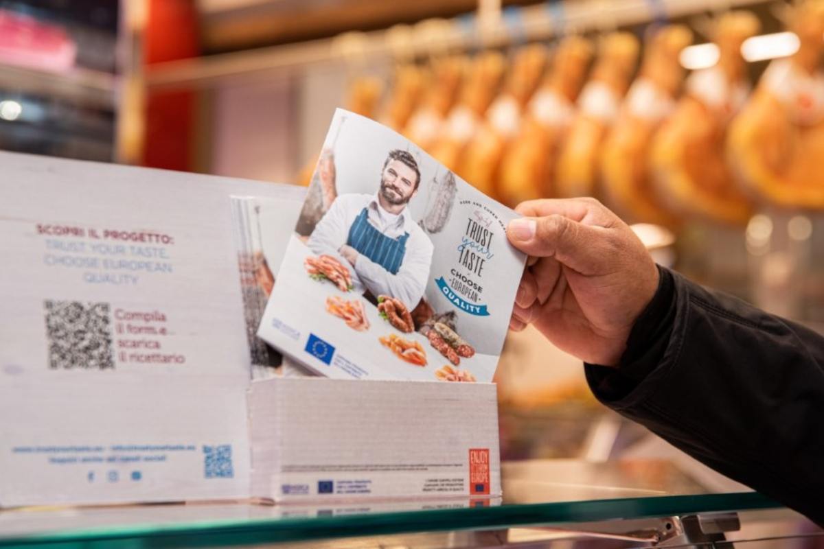 Mano tiene volantino Assica su carne suina italiana in punto vendita con prosciutti appesi