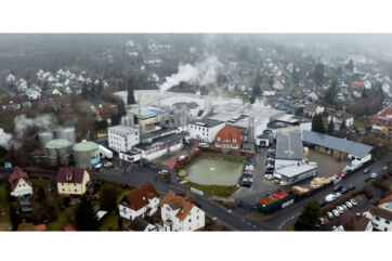 Aerial view of Immergut factory, a food processing plant, with smoke, silos, buildings, and a pond in a residential area.