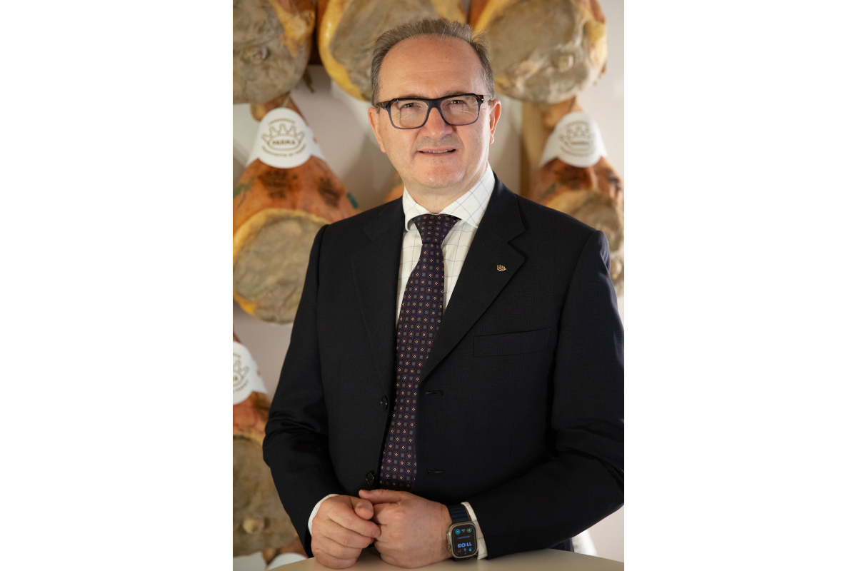 Alessandro Utini, Presidente del Consorzio del Prosciutto di Parma, in completo, con il prosciutto di Parma sullo sfondo.