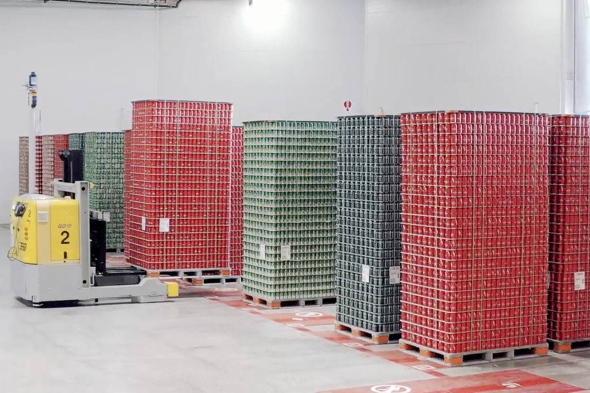 Automated guided vehicle (AGV) transports pallets of canned goods in a warehouse. Block storage of red, green, and gray cans.