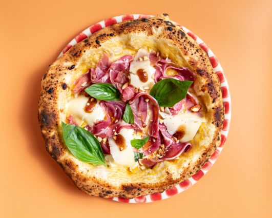 Giuseppe Cutraro's pizza, from Paris. Gourmet pizza with basil, prosciutto, cheese, and balsamic glaze on a red and white plate.