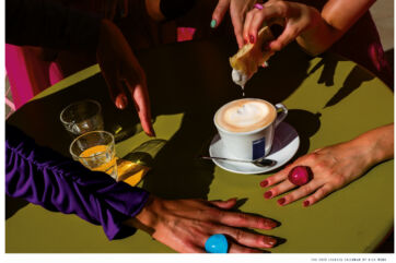 Lavazza coffee cup with latte art on a green table. Hands with colorful rings and manicured nails surround the coffee and pastries.