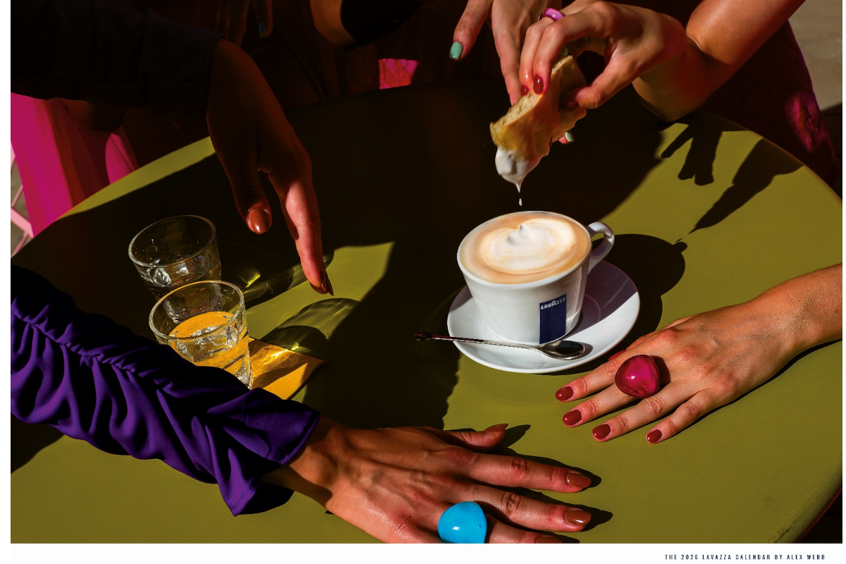 Lavazza coffee cup with latte art on a green table. Hands with colorful rings and manicured nails surround the coffee and pastries.