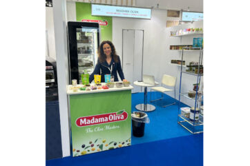 Madama Oliva booth at Foodex Japan. Woman stands behind olive product display. Shelves with olive jars and snacks.