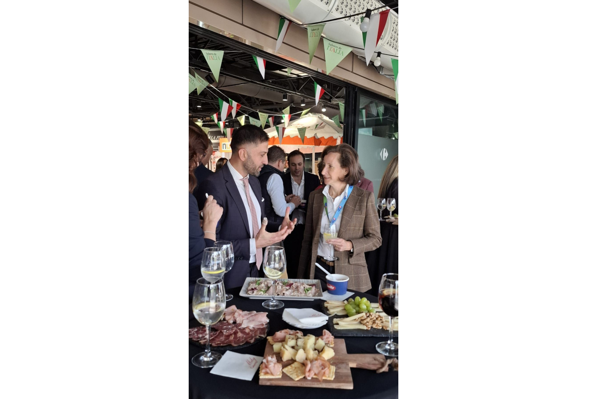 People at Italian Week event at Carrefour Spain. Food and drinks on table. Italian flags hanging.