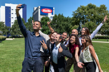 Barilla team selfie. Happy employees celebrate Barilla's top food company ranking, outdoors with Barilla HQ in background.