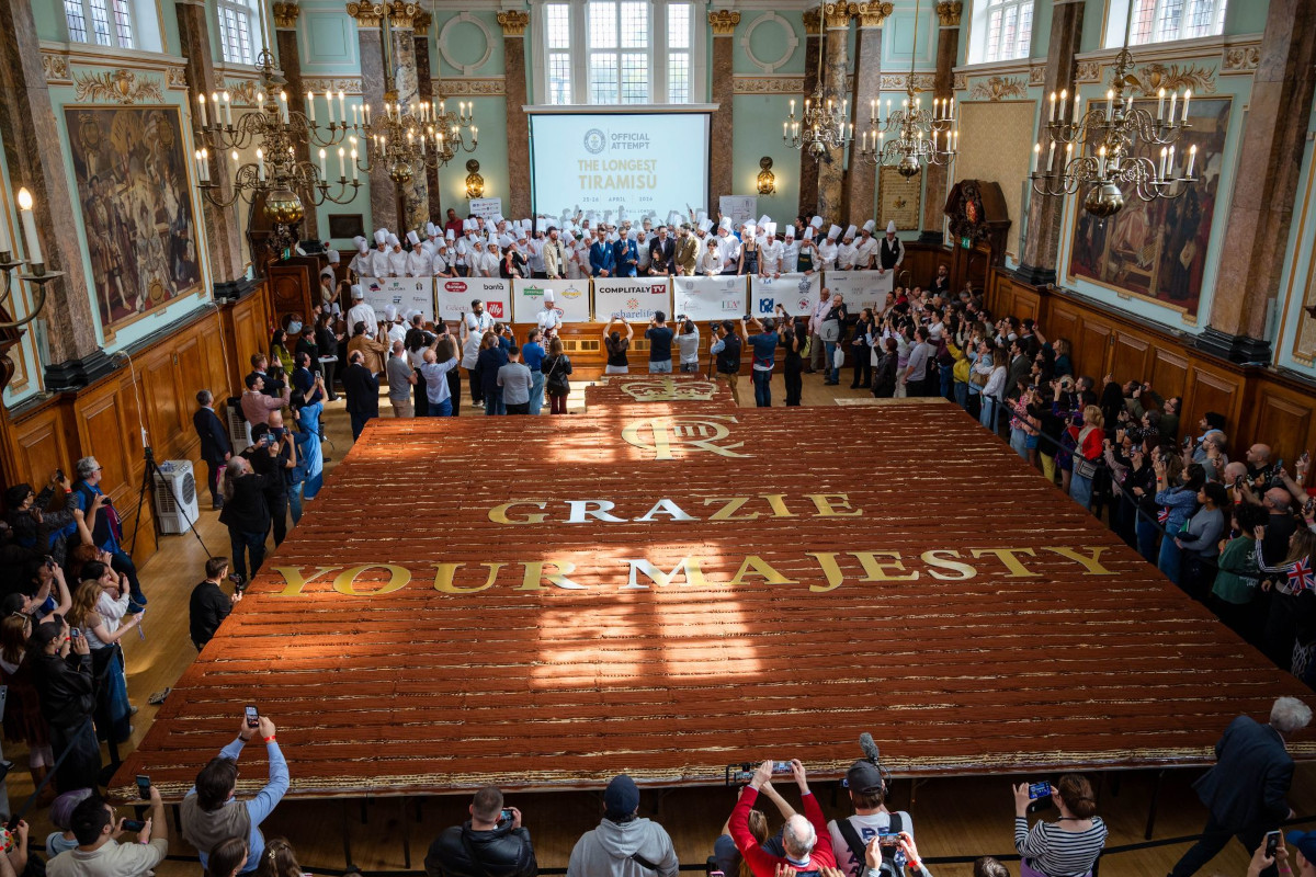 Crowd views world's longest tiramisu. Grazie Your Majesty written on dessert. Chefs, onlookers, and cameras.