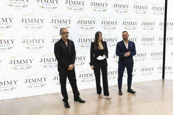 Jimmy Tartufi event. Three people stand in front of a branded backdrop. Fashionable attire, possible product launch.