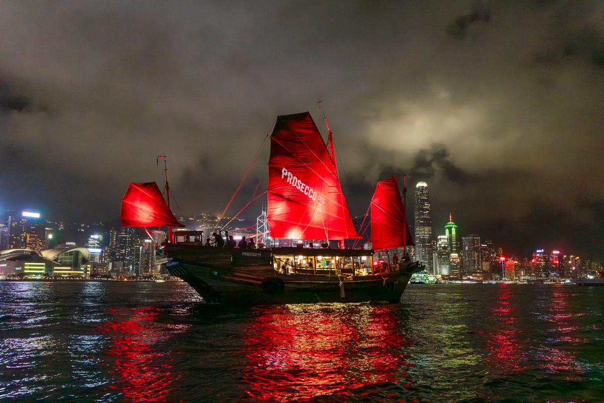 Hong Kong skyline at night with a red-sailed junk boat in the harbor. Vinexpo Hong Kong.