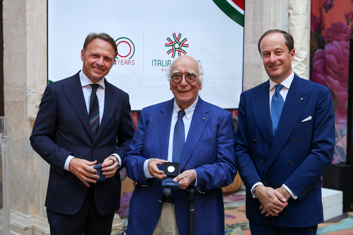 Three men in suits at an Italia Food event, with the center man holding a commemorative coin in a presentation box.