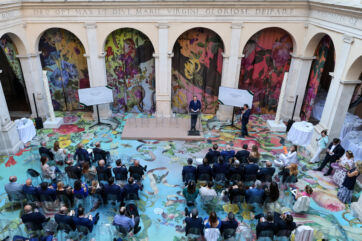 Speaker at a corporate event in a historic courtyard with floral floor decor, large screens, and an audience seated. - Italia del Gusto