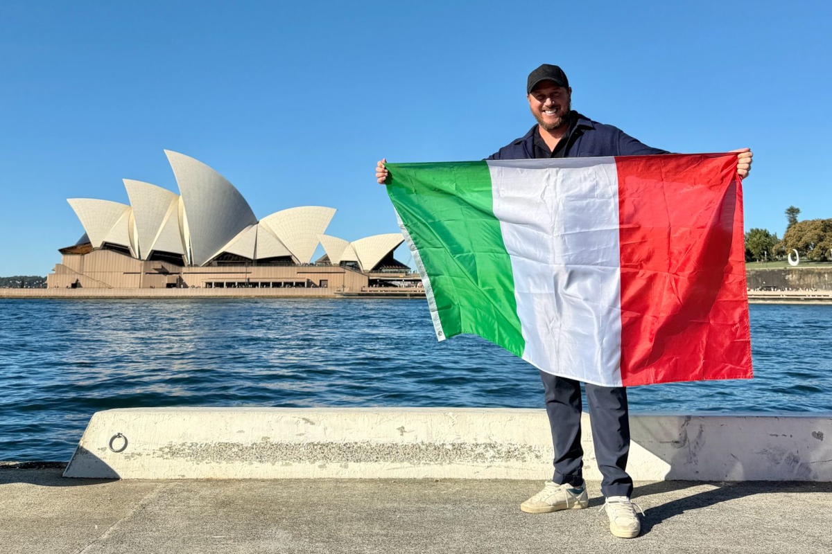 Smiling man holding an Italian flag in front of the Sydney Opera House on a sunny day in Australia.
