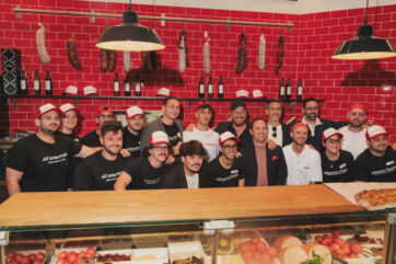 Group of All'Antico Vinaio staff in branded caps and shirts standing behind a deli counter with hanging cured meats.