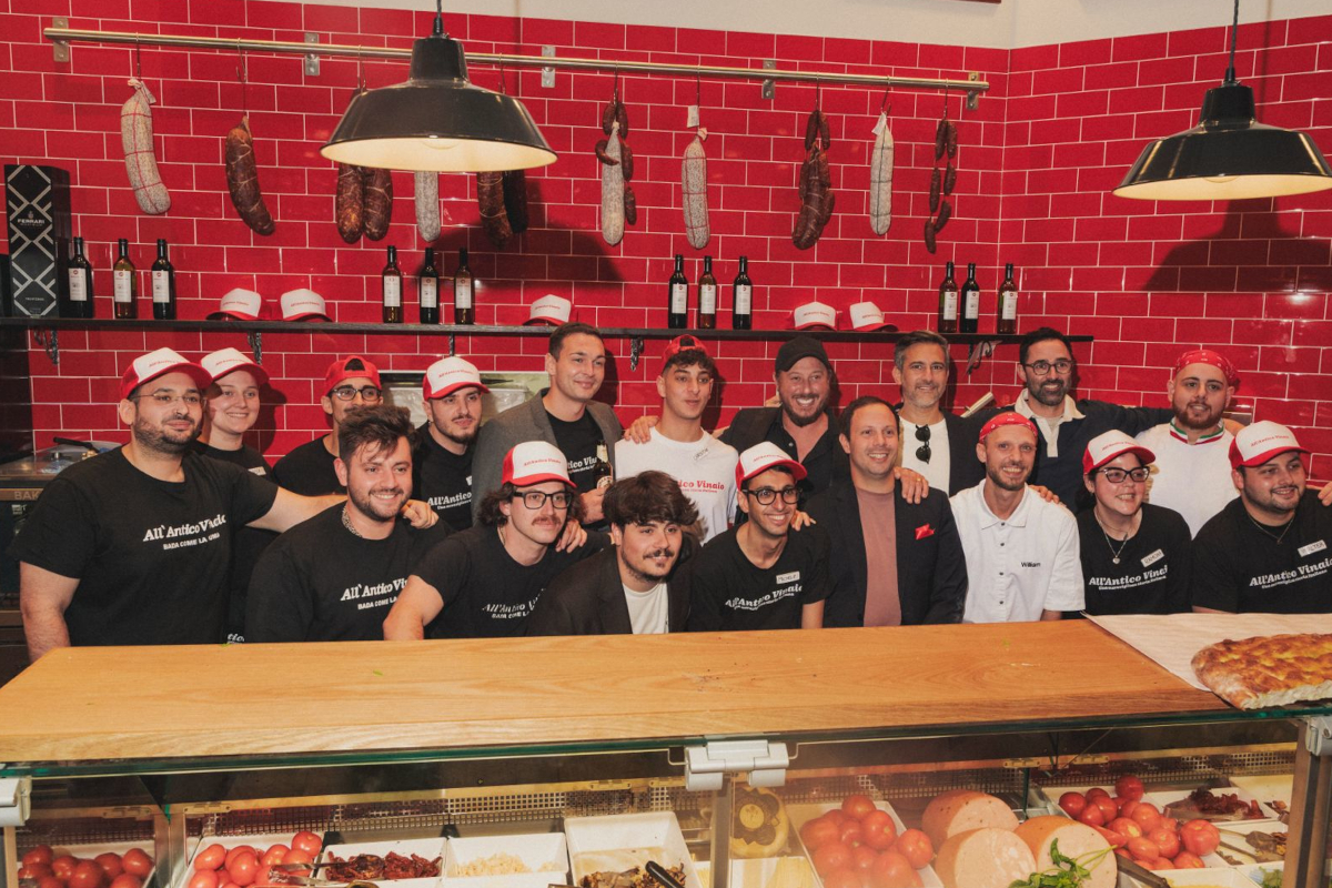 Group of All'Antico Vinaio staff in branded caps and shirts standing behind a deli counter with hanging cured meats.