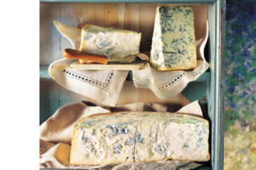 Gorgonzola cheese wedges and a large wheel displayed on vintage linen napkins inside a rustic blue wooden credenza shelf.