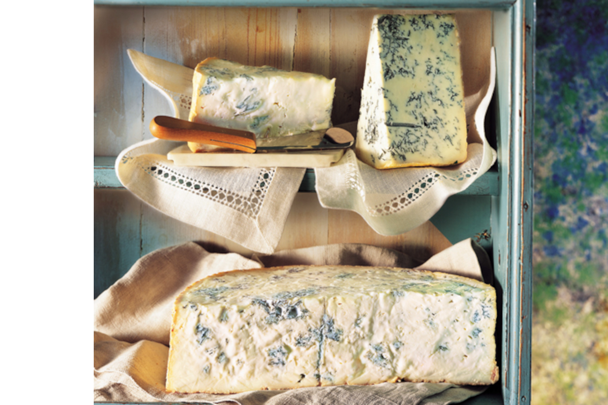 Gorgonzola cheese wedges and a large wheel displayed on vintage linen napkins inside a rustic blue wooden credenza shelf.