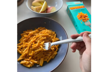 A hand using a fork to eat a bowl of Goodles mac and cheese, with the branded box and a bowl of apple slices nearby.