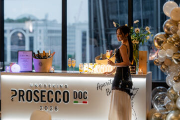 Woman holding a Prosecco DOC cocktail at a branded event bar with ice buckets, glasses, and gold balloon decorations.