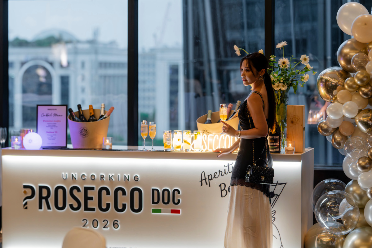 Woman holding a Prosecco DOC cocktail at a branded event bar with ice buckets, glasses, and gold balloon decorations.