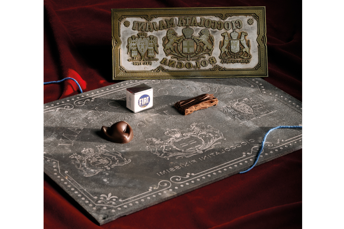 Vintage Majani chocolate packaging: historical tin sign, antique chocolate box, clay pipe and cacao beans on grey cloth.