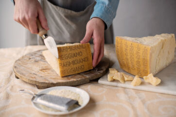 Hands slicing a wheel of Parmigiano Reggiano PDO cheese with a knife at SIAL Paris, showing crumbles and a grater.