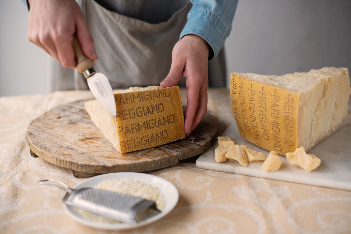 Hands slicing a wheel of Parmigiano Reggiano PDO cheese with a knife at SIAL Paris, showing crumbles and a grater.