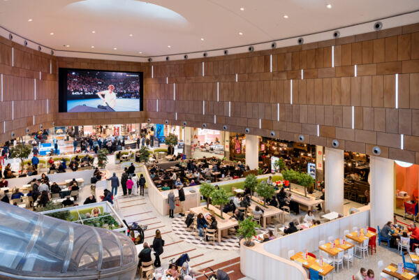 Area food court affollata. Ristorazione varia, design moderno, schermi TV. Persone sedute ai tavoli.